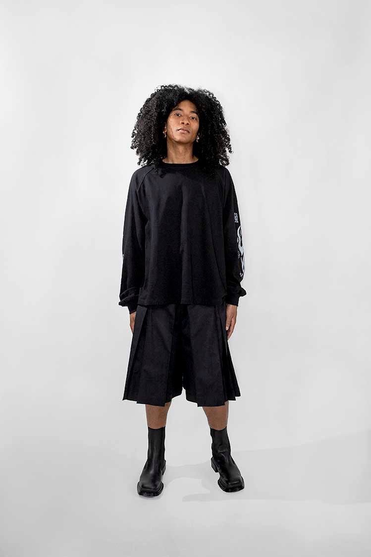 A model with curly black hair stands against a light gray studio background wearing a black long-sleeve top, black pleated shorts, and black ankle boots.