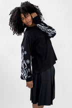 A model with voluminous curly black hair wearing a black long-sleeve top with white flame and floral patterns on the sleeves, paired with a black pleated skort, against a light gray studio background.