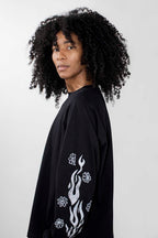A person with voluminous curly black hair wearing an oversized black long-sleeve shirt with white flame and flower graphics on the sleeve, photographed against a light gray studio background.