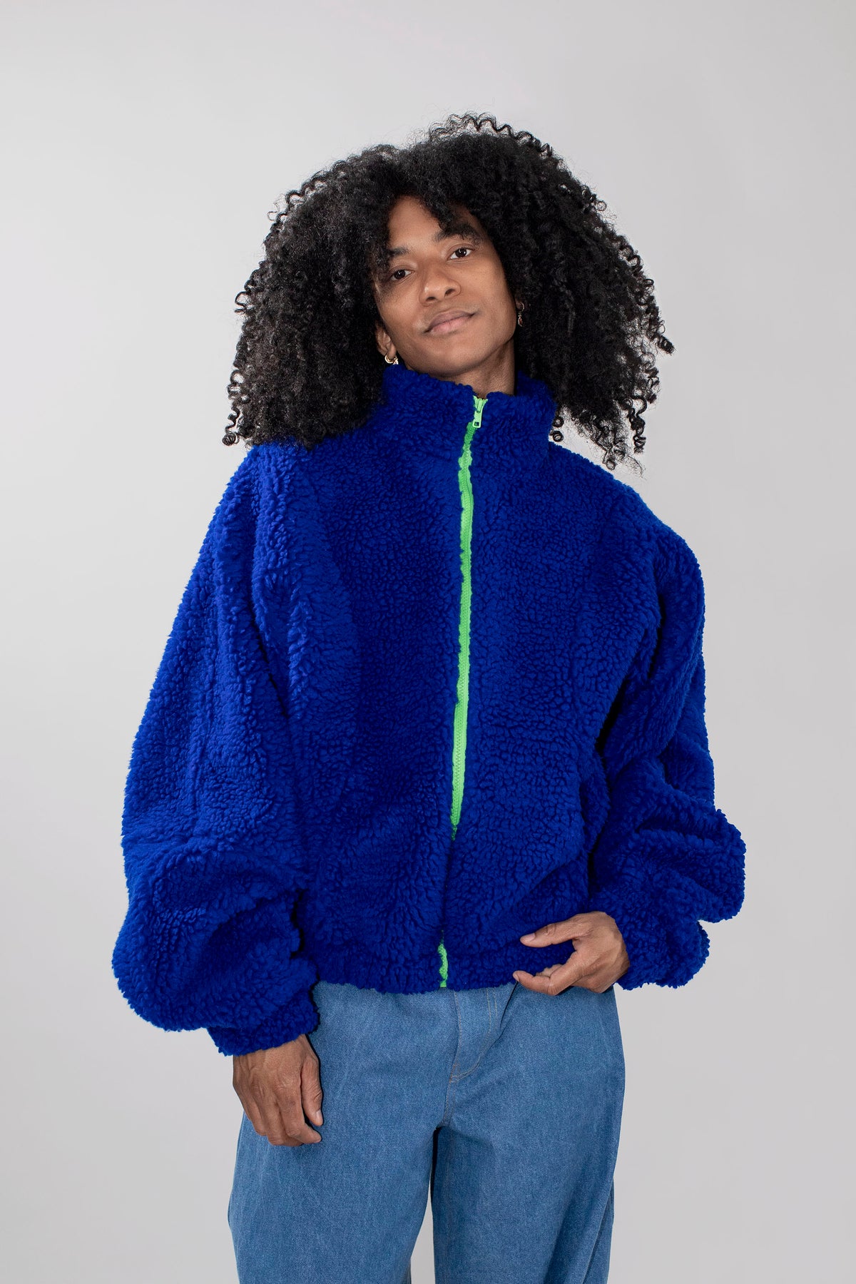 A person with curly black hair wearing a bright royal blue teddy fleece zip-up jacket with a neon green zipper, paired with light blue jeans, against a plain light gray studio background.