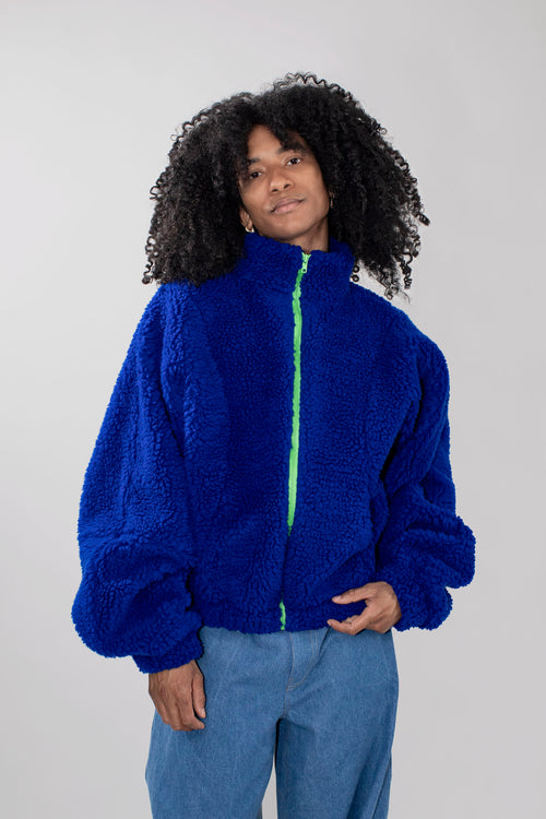 A person with curly black hair wearing a bright royal blue teddy fleece zip-up jacket with a neon green zipper, paired with light blue jeans, against a plain light gray studio background.