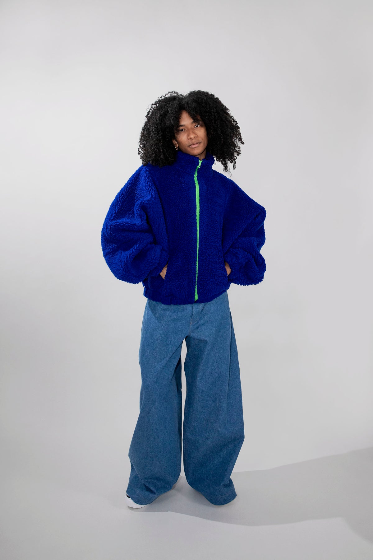 A model with curly black hair stands against a light gray studio background wearing a bright royal blue fuzzy zip-up jacket with a neon green zipper and wide-leg blue pants.