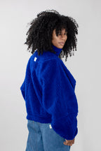 A model with curly black hair is shown from a side-back angle wearing a bright royal blue teddy fleece zipper jacket and blue jeans against a plain light gray studio background.