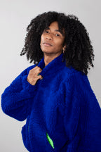 A person with voluminous black curly hair wearing a bright royal blue fuzzy fleece jacket, posing against a light gray studio background.