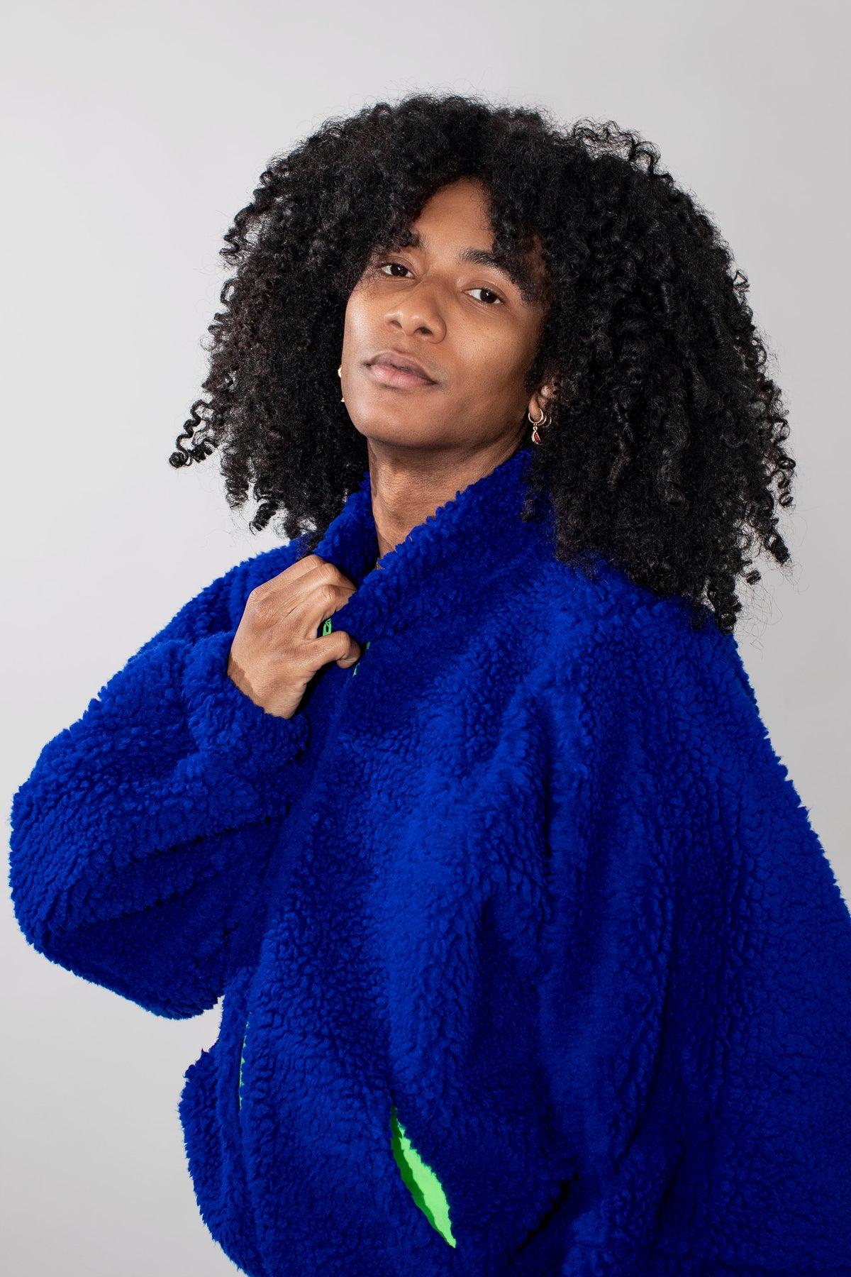 A person with voluminous black curly hair wearing a bright royal blue fuzzy fleece jacket, posing against a light gray studio background.