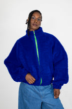 A model wearing a vibrant royal blue teddy fleece zip-up jacket with a contrasting neon green zipper, paired with light blue jeans, against a plain white studio background.