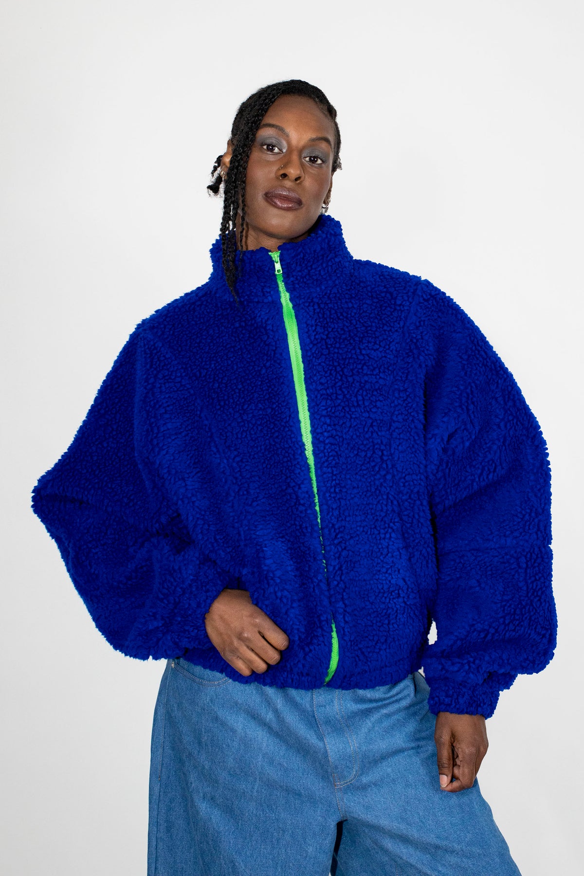 A model wearing a vibrant royal blue teddy fleece zip-up jacket with a contrasting neon green zipper, paired with light blue jeans, against a plain white studio background.