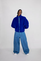 A model stands against a light gray studio background wearing a bright royal blue fuzzy zip-up jacket with a neon green zipper line and oversized blue jeans.