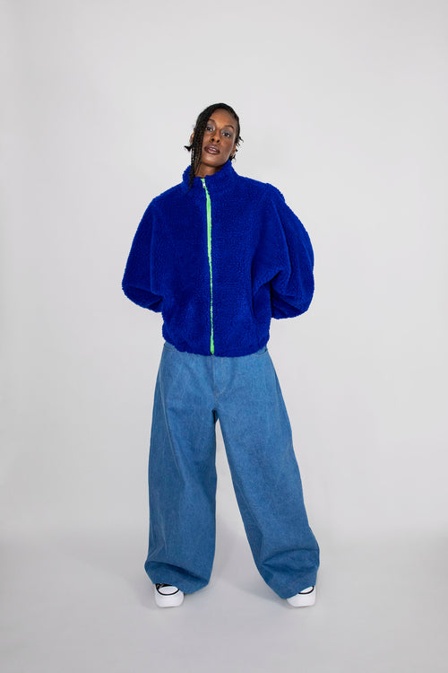 A model stands against a light gray studio background wearing a bright royal blue fuzzy zip-up jacket with a neon green zipper line and oversized blue jeans.
