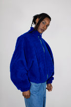 A model wearing a vibrant royal blue teddy fleece zip-up jacket with a high collar and light blue jeans, photographed against a neutral light gray studio background.