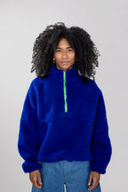 A model with curly black hair wearing a bright royal blue teddy fleece half zip pullover with a neon green zipper, paired with light blue jeans, against a solid light gray background.