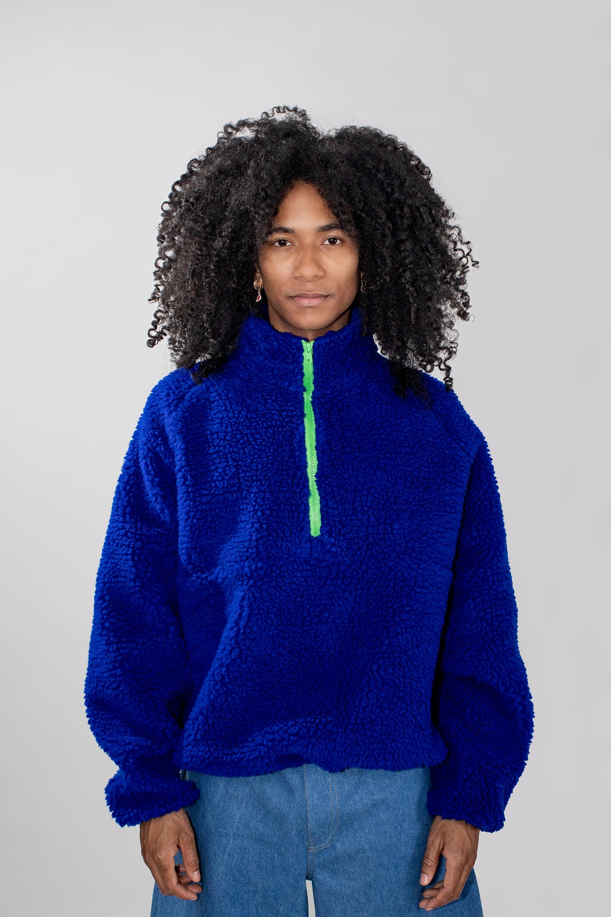 A model with curly black hair wearing a bright royal blue teddy fleece half zip pullover with a neon green zipper, paired with light blue jeans, against a solid light gray background.