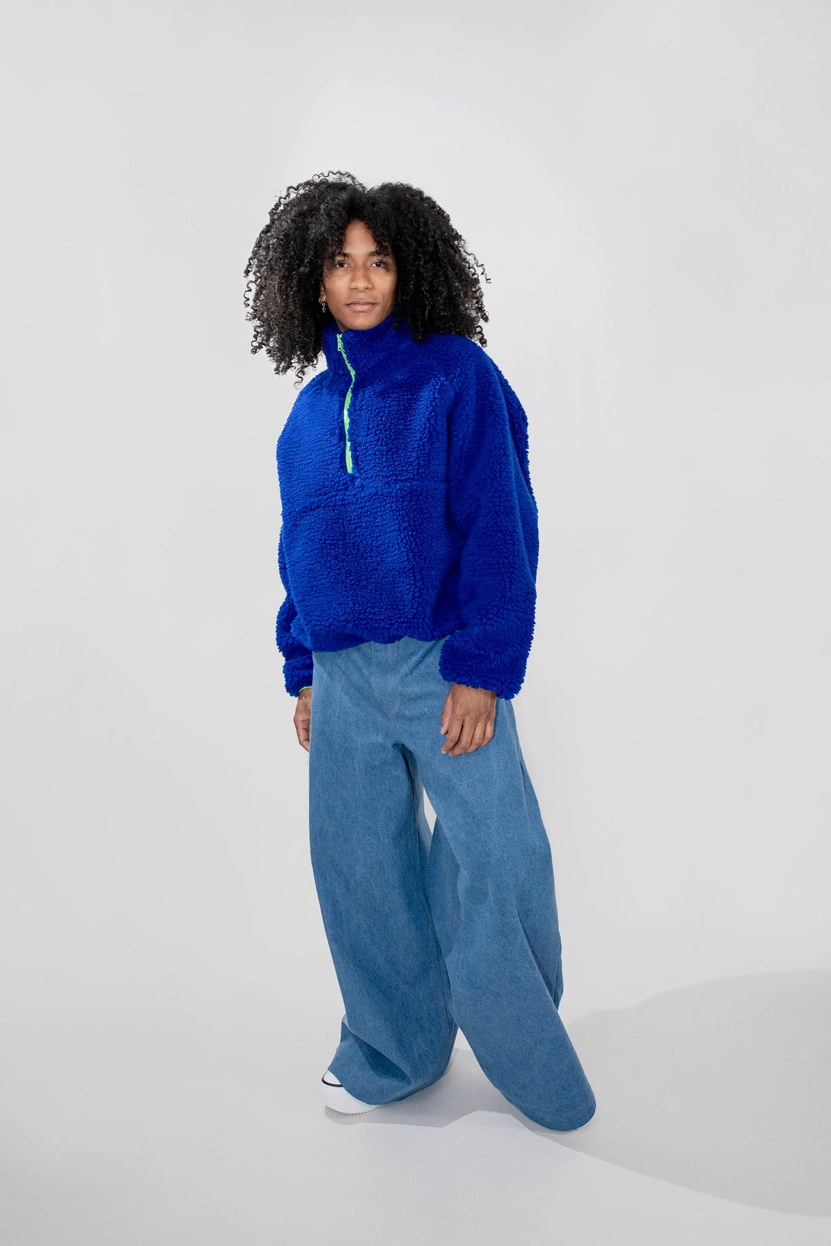A model with curly black hair stands against a light gray studio background wearing a bright royal blue fuzzy half zip pullover and wide-leg blue jeans.