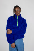 A model wearing a bright royal blue teddy fleece half zip with a neon green zipper, paired with light blue jeans, against a plain light gray background.