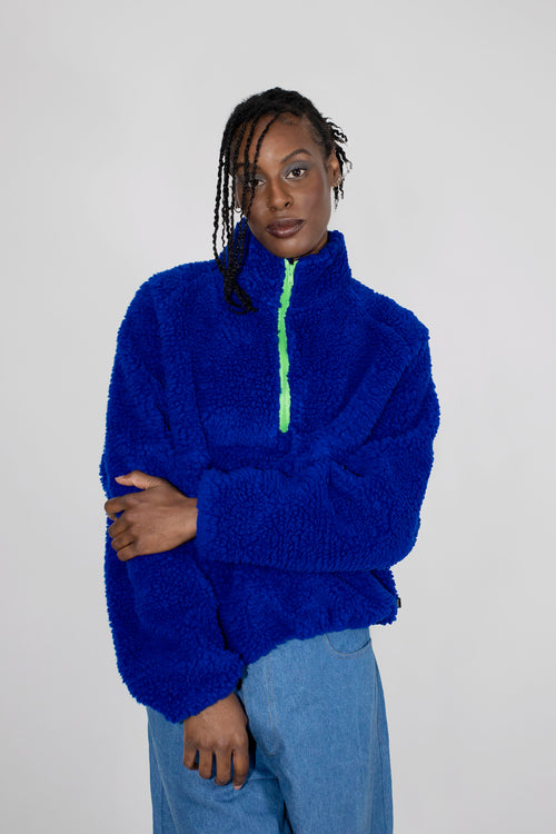 A model wearing a bright royal blue teddy fleece half zip with a neon green zipper, paired with light blue jeans, against a plain light gray background.