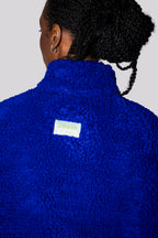 Close-up back view of a person wearing a bright royal blue teddy fleece pullover jacket, with a small white brand label reading 'blk top KOPE New York City' visible at the collar.