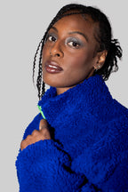 A close-up portrait of a woman wearing a vibrant royal blue fuzzy fleece jacket against a solid light gray background.