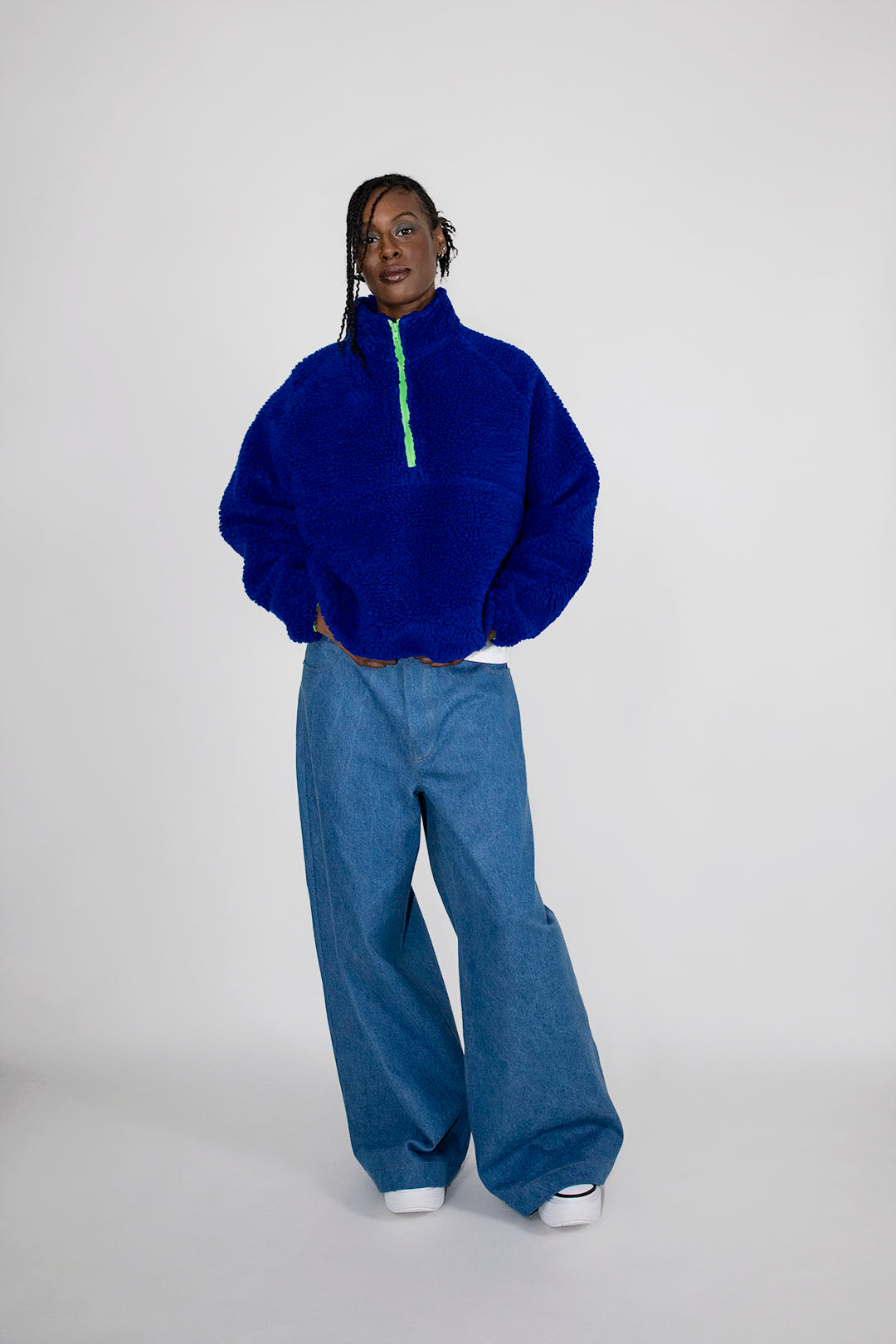 A model wearing a vibrant royal blue teddy fleece half-zip pullover with a neon green zipper, paired with wide-leg blue jeans and white sneakers, standing against a light gray studio background.