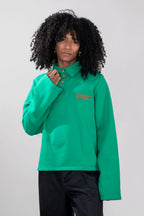 A model wearing a green long-sleeve polo and loose black pants, standing against a light gray studio background.