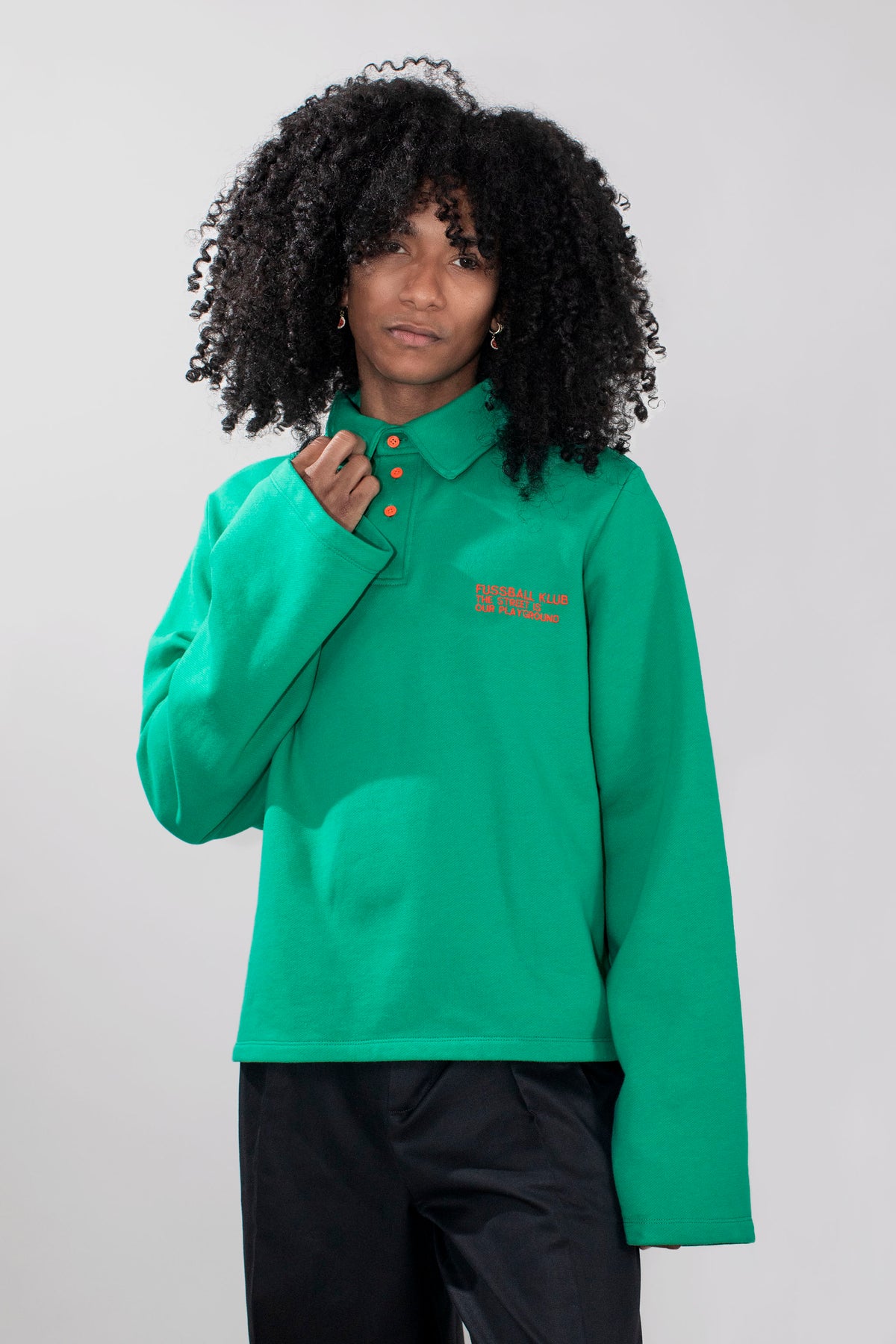 A model wearing a green long-sleeve polo and loose black pants, standing against a light gray studio background.