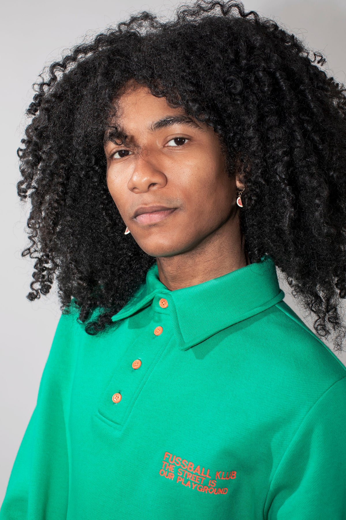 A model with curly black hair wearing a green long-sleeve polo shirt with neon orange buttons and neon orange embroidered text on the chest, against a solid light gray background.