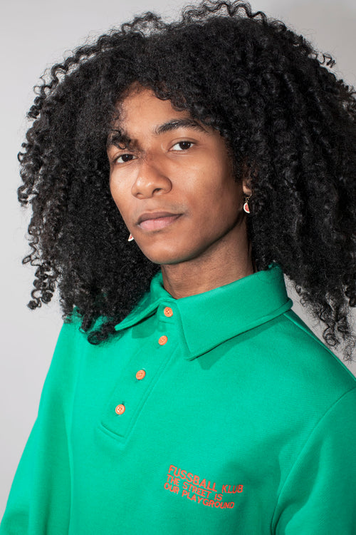 A model with curly black hair wearing a green long-sleeve polo shirt with neon orange buttons and neon orange embroidered text on the chest, against a solid light gray background.