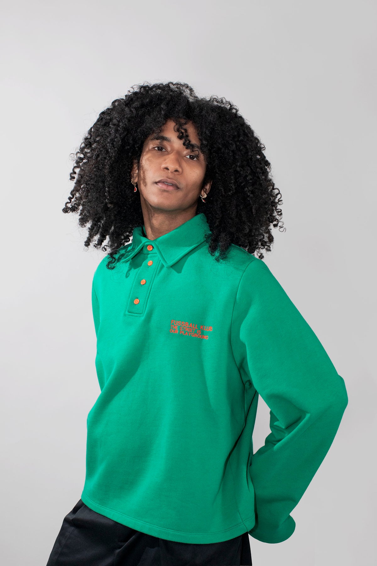 A model with curly black hair wearing a green long-sleeve polo shirt with neon orange buttons and neon orange embroidered text on the chest, paired with black pants, against a solid light gray background.