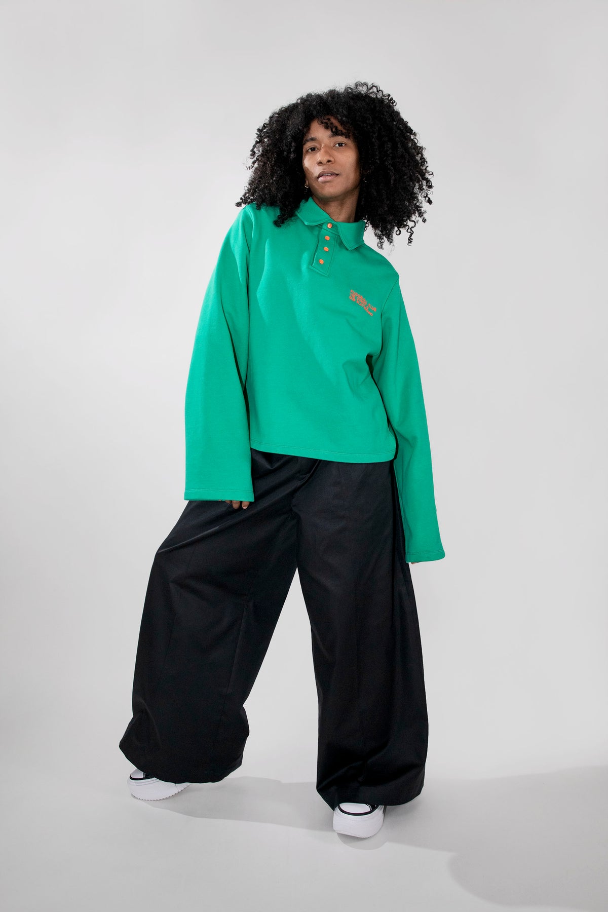A model with curly black hair stands against a light gray studio background wearing a green long-sleeve polo shirt, black wide-leg pants, and white sneakers.