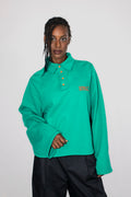 A model wearing a green long-sleeve polo shirt with neon orange buttons and neon orange embroidery, paired with black pants, against a light gray studio background.