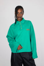 A model wearing a green long-sleeve polo shirt with neon orange buttons and neon orange embroidery, paired with black pants, against a light gray studio background.