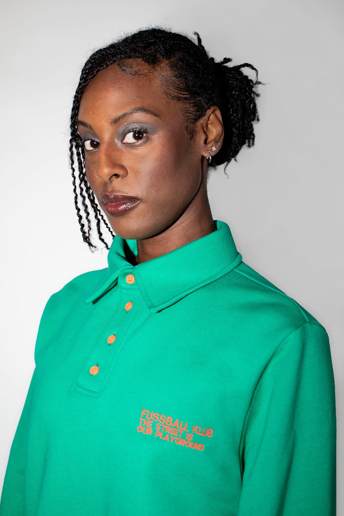 A woman with braided hair wearing a green polo shirt with neon orange buttons and neon orange embroidered text on the left chest, photographed against a plain light gray background.