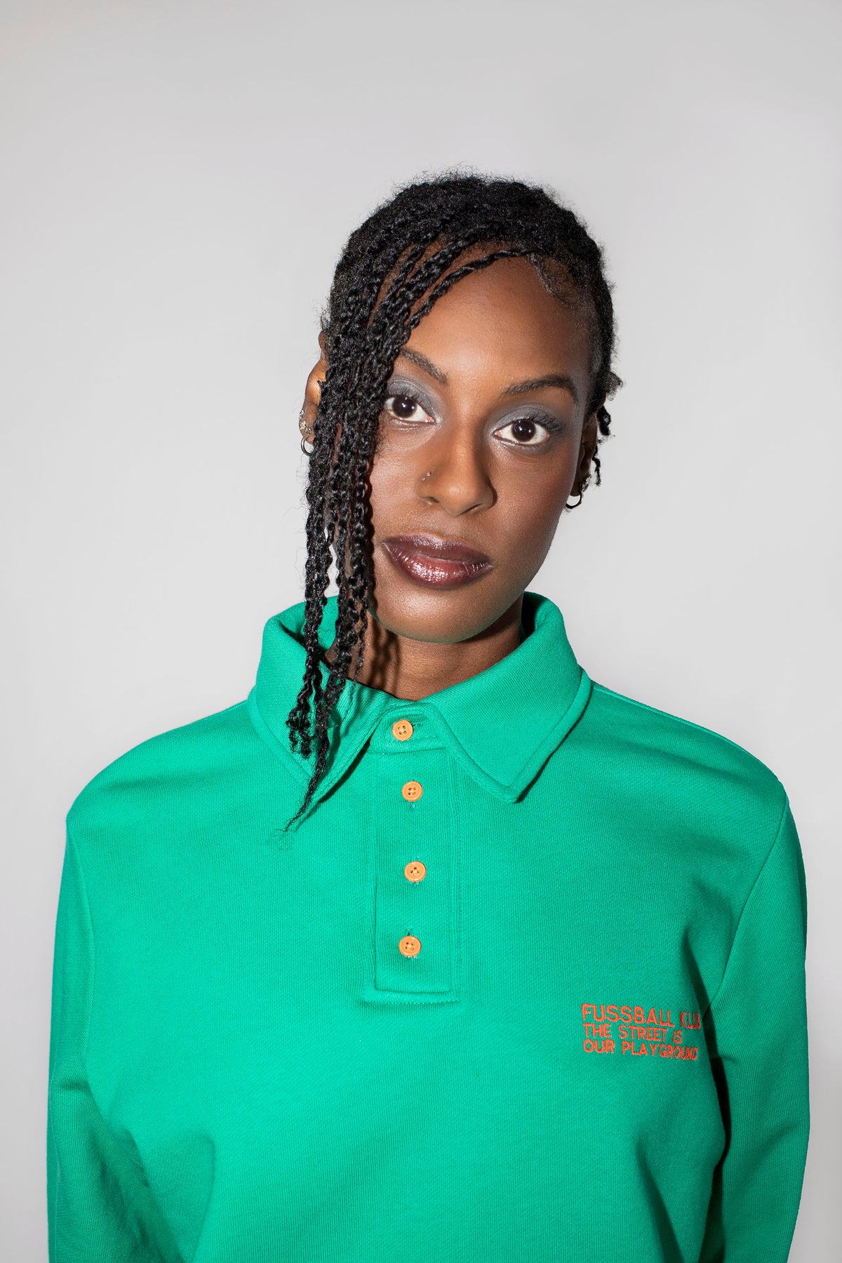 A woman with braided hair wearing a green long-sleeve polo shirt with neon orange buttons and neon orange embroidered text on the chest, photographed against a plain light gray background.