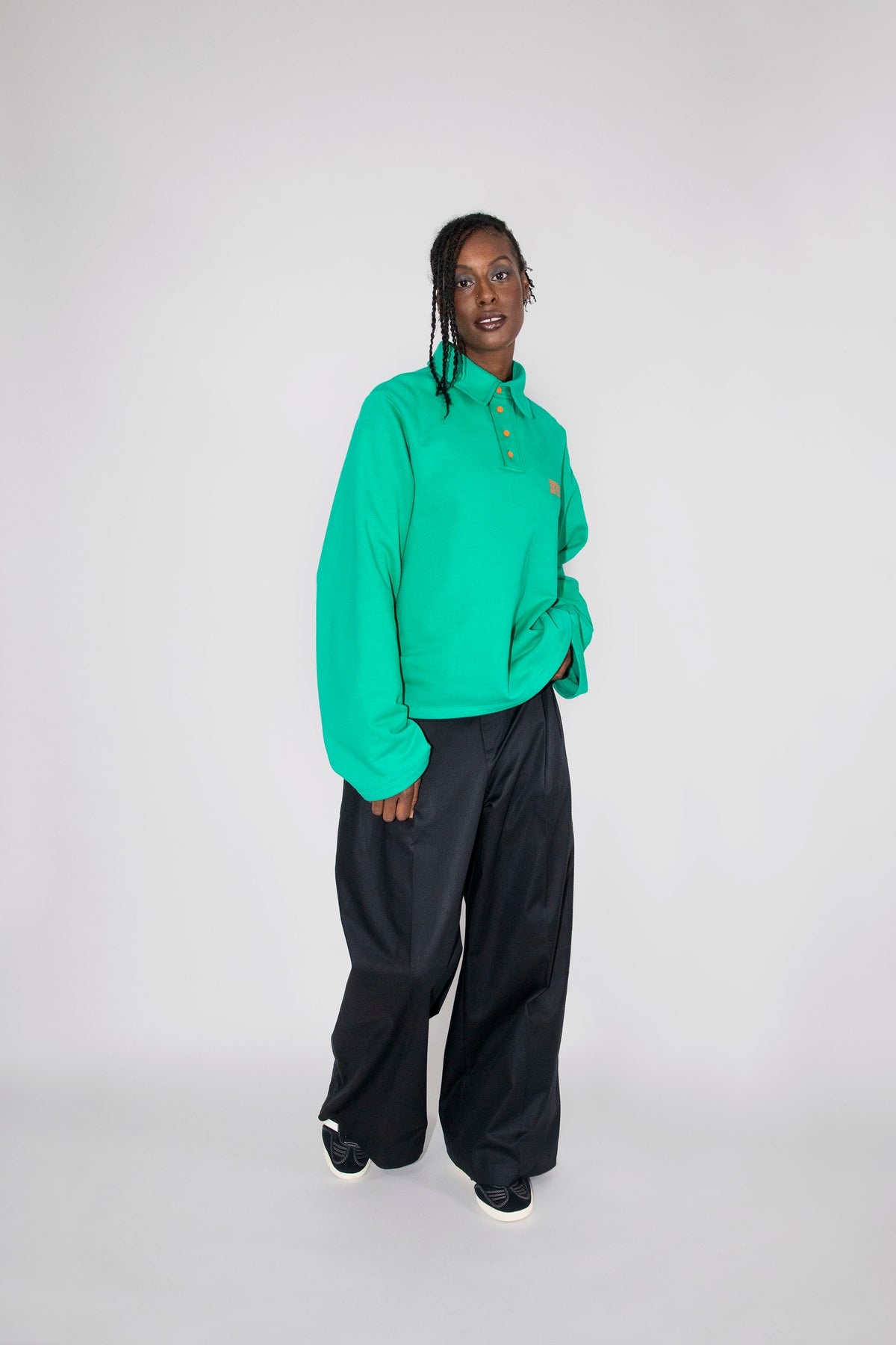 A model wearing a green long-sleeve polo and loose black pants, standing against a light gray studio background.
