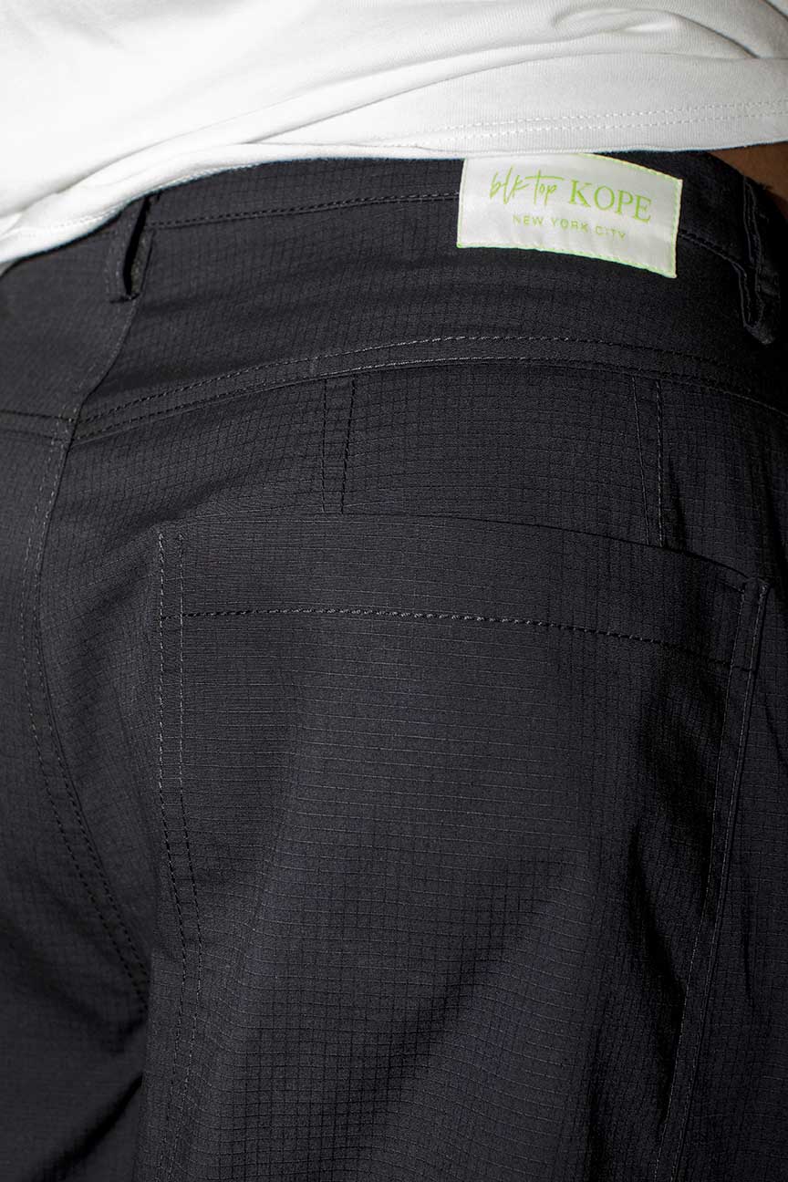 Close-up of the back waistband and pocket area of dark charcoal pants, with a white shirt hem visible at the top and a small light label reading blk top KOPE.
