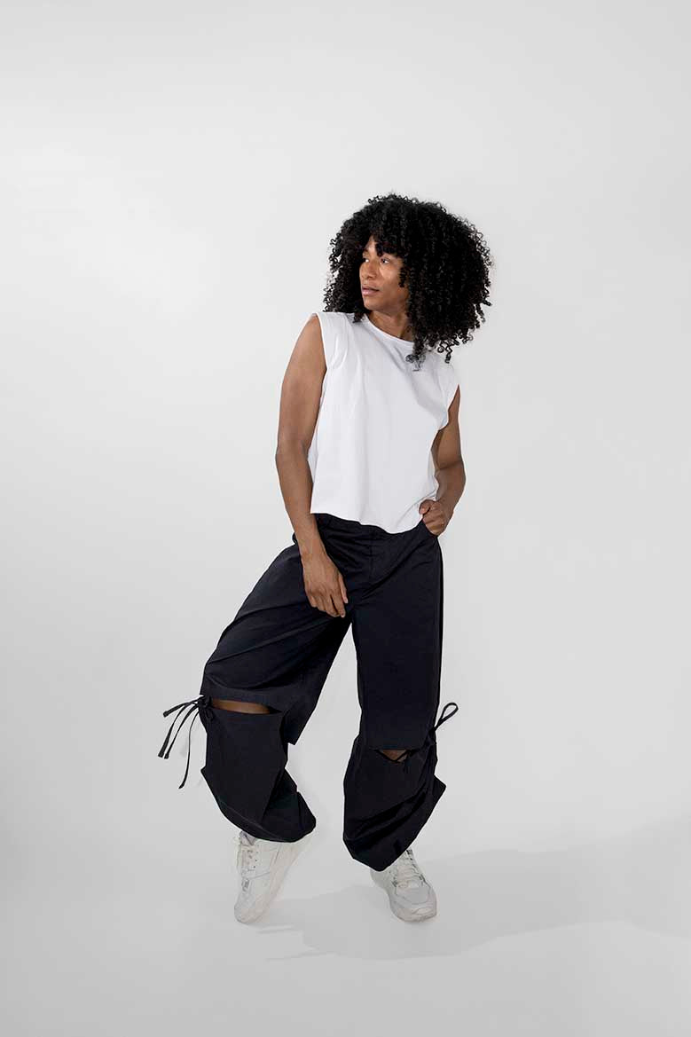 A model with curly black hair is wearing a sleeveless white top, loose black pants with drawstring details, and white sneakers, posing against a light gray studio background.