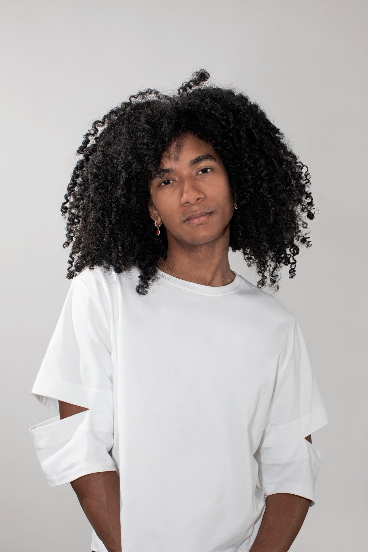 A person with black curly hair wearing a white oversized T-shirt with cutout details on the sleeves, photographed against a plain light gray studio background.