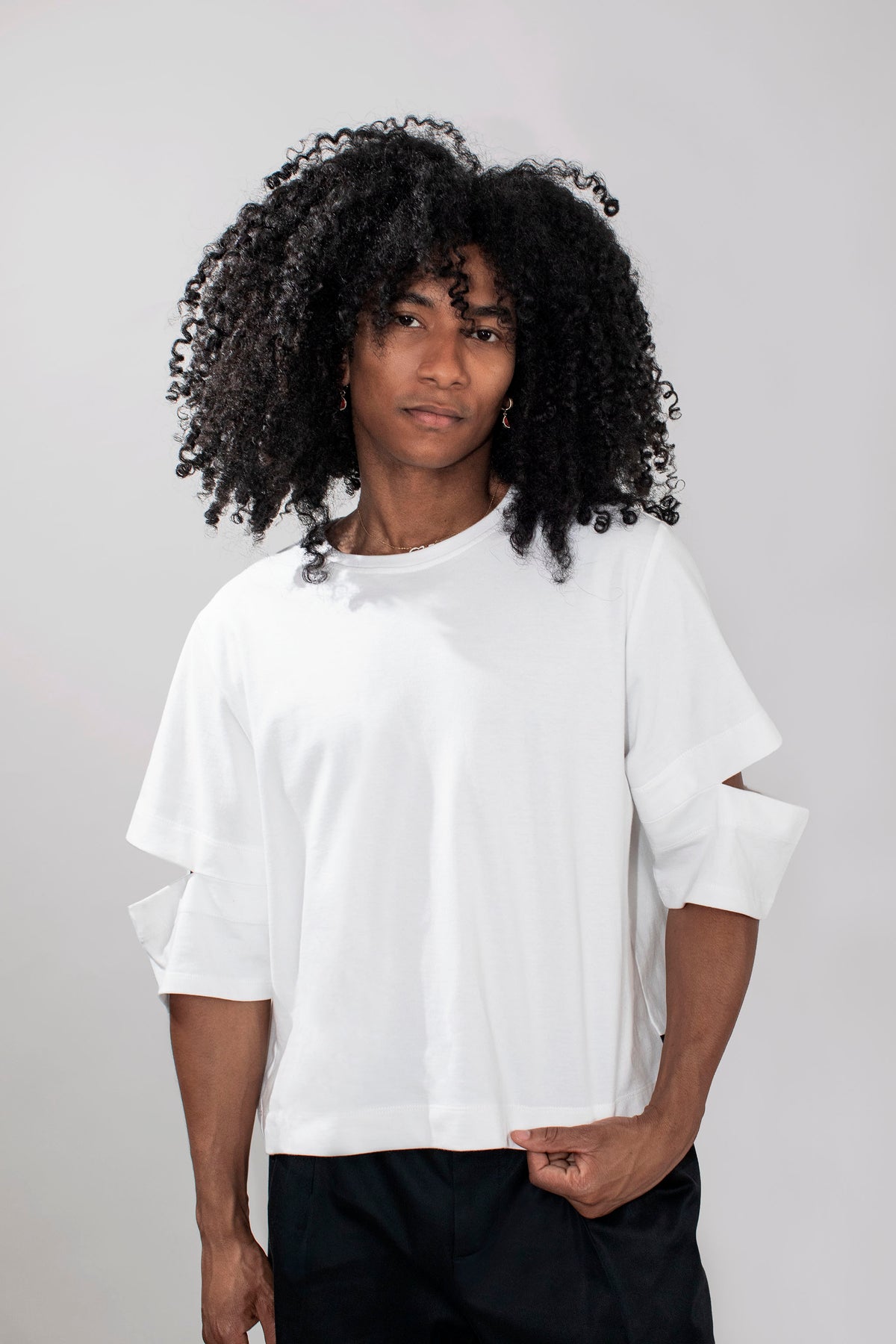 A model with voluminous black curly hair wearing a white oversized short-sleeve t-shirt with cutout details on the sleeves and black pants, standing against a light gray studio background.