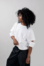 A model with curly black hair wearing a white oversized short-sleeve t-shirt with cutout details on the sleeves and black wide-leg pants, photographed against a neutral light gray background.