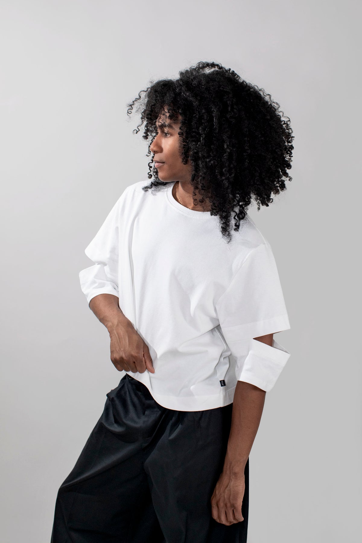 A model with curly black hair wearing a white oversized short-sleeve t-shirt with cutout details on the sleeves and black wide-leg pants, photographed against a neutral light gray background.