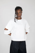 A model wearing a white short-sleeve crewneck sweatshirt with cutout details on the sleeves, paired with black trousers, against a light gray studio background.