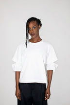 A model wearing a white oversized short-sleeve t-shirt with cutout details on the sleeves, paired with black trousers, against a plain light gray background.