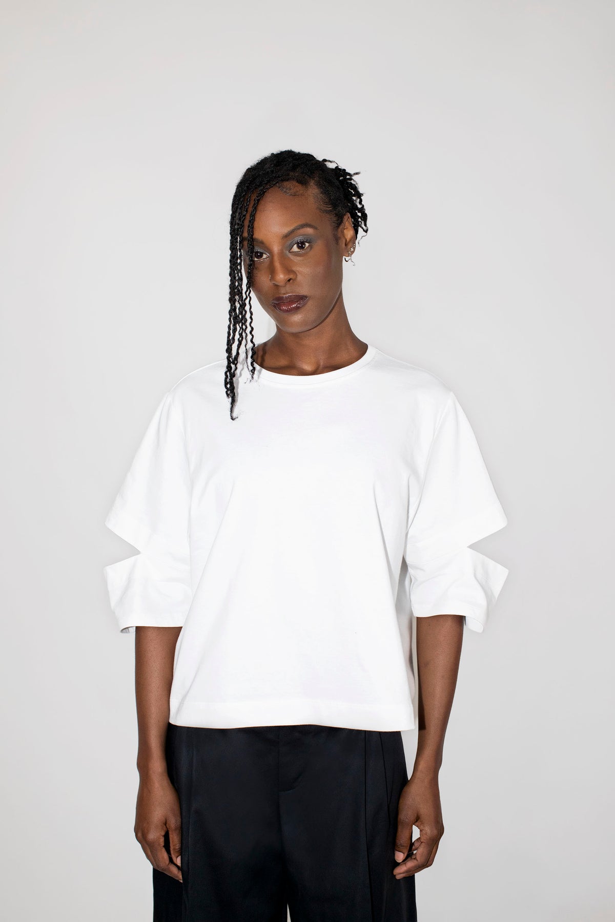 A model wearing a white oversized short-sleeve t-shirt with cutout details on the sleeves, paired with black trousers, against a plain light gray background.