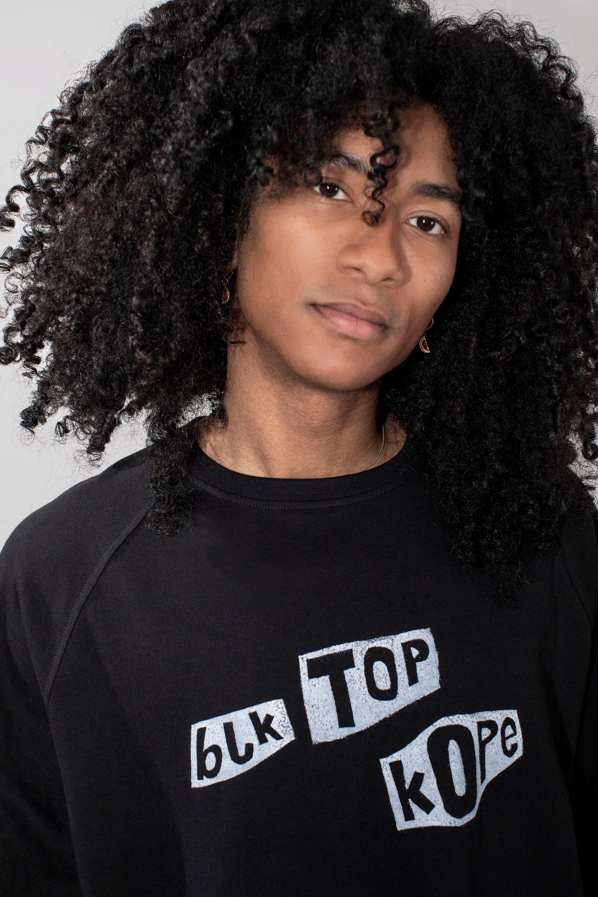 A person with voluminous curly black hair wearing a black long-sleeve sweatshirt with a white graphic text print that reads 'blk TOP kOpe'.