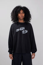 A model with curly black hair wearing a black long-sleeve crewneck sweatshirt with a white graphic text print reading 'BLK TOP KOPE' on the chest, paired with black pants, against a light gray studio background.