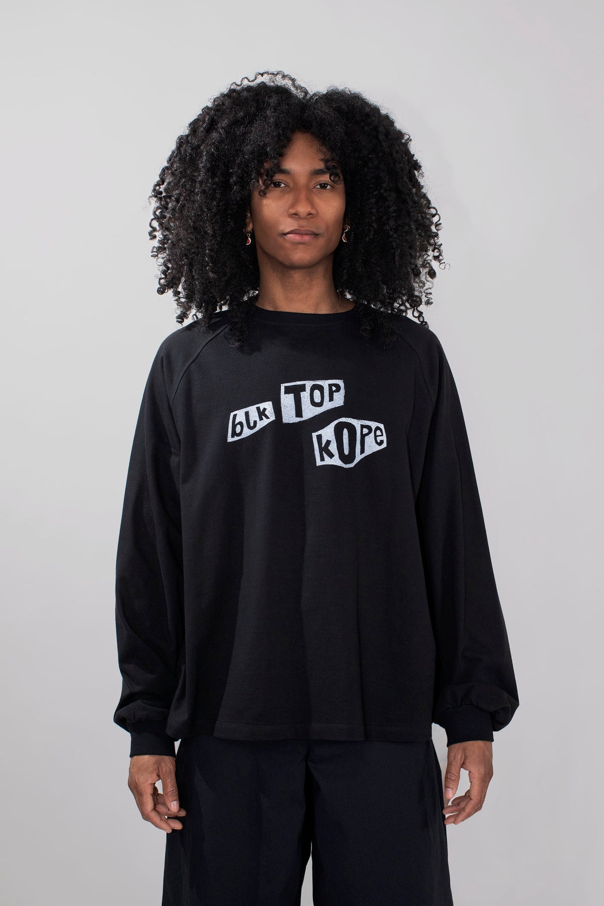 A model with curly black hair wearing a black long-sleeve crewneck sweatshirt with a white graphic text print reading 'BLK TOP KOPE' on the chest, paired with black pants, against a light gray studio background.