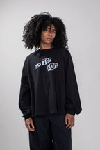 A model with curly black hair wearing a black long-sleeve sweatshirt with a white graphic text print on the chest, paired with black pants, against a plain light gray background.