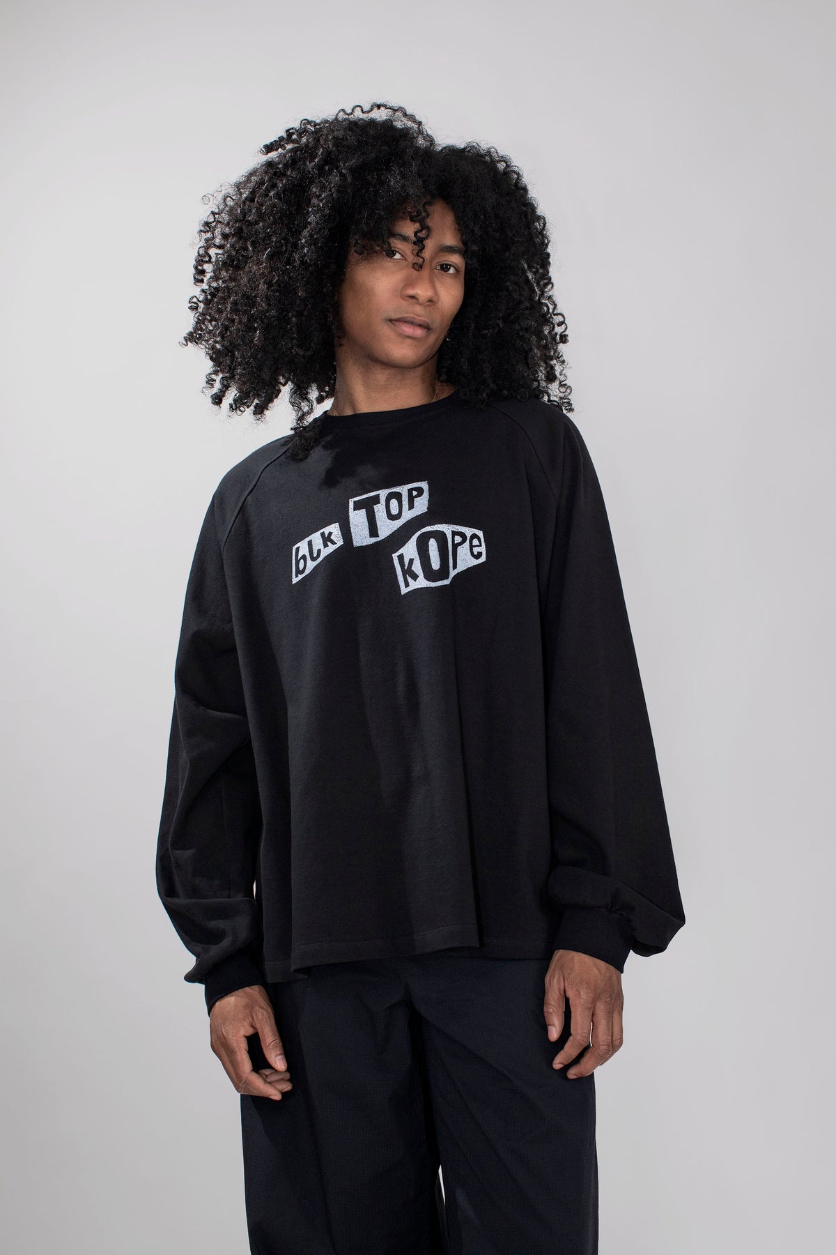 A model with curly black hair wearing a black long-sleeve sweatshirt with a white graphic text print on the chest, paired with black pants, against a plain light gray background.