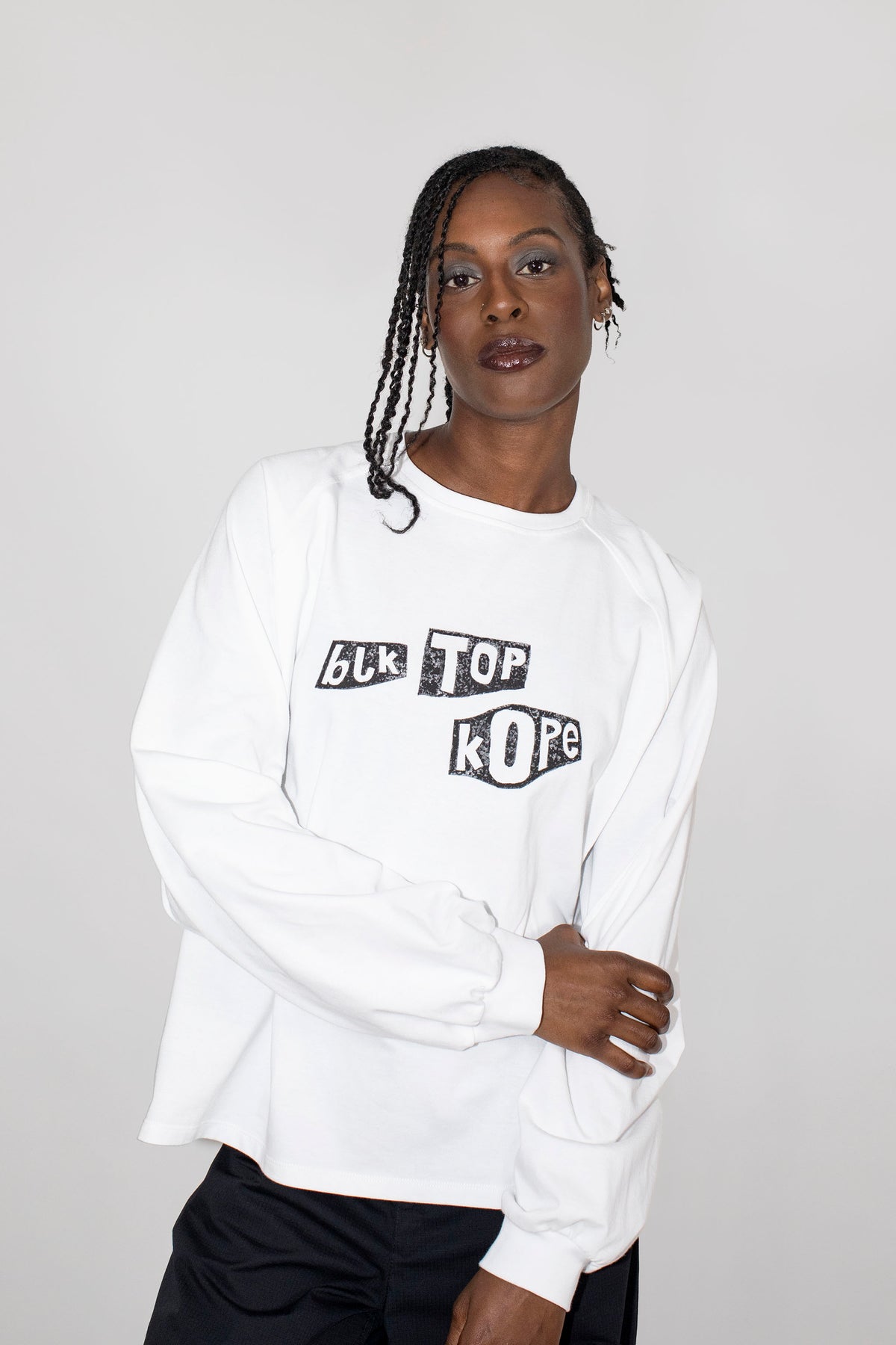 A model wearing a white long-sleeve sweatshirt with a black graphic text print reading 'BLK TOP KOPE' on the chest, paired with black pants, against a light gray studio background.