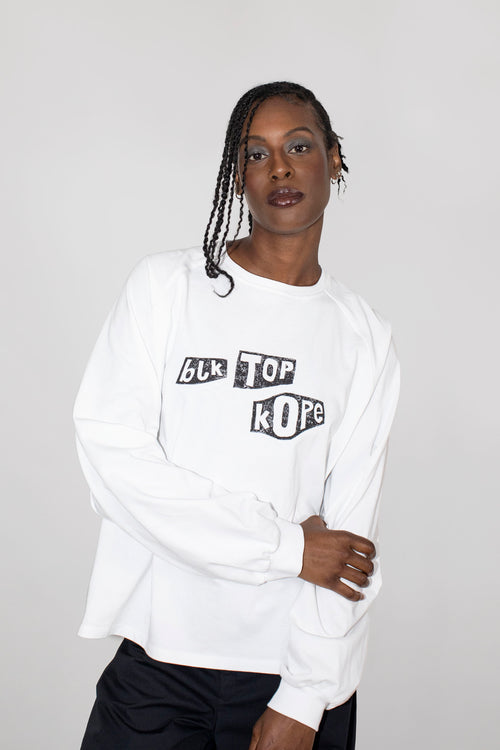 A model wearing a white long-sleeve sweatshirt with a black graphic text print reading 'BLK TOP KOPE' on the chest, paired with black pants, against a light gray studio background.