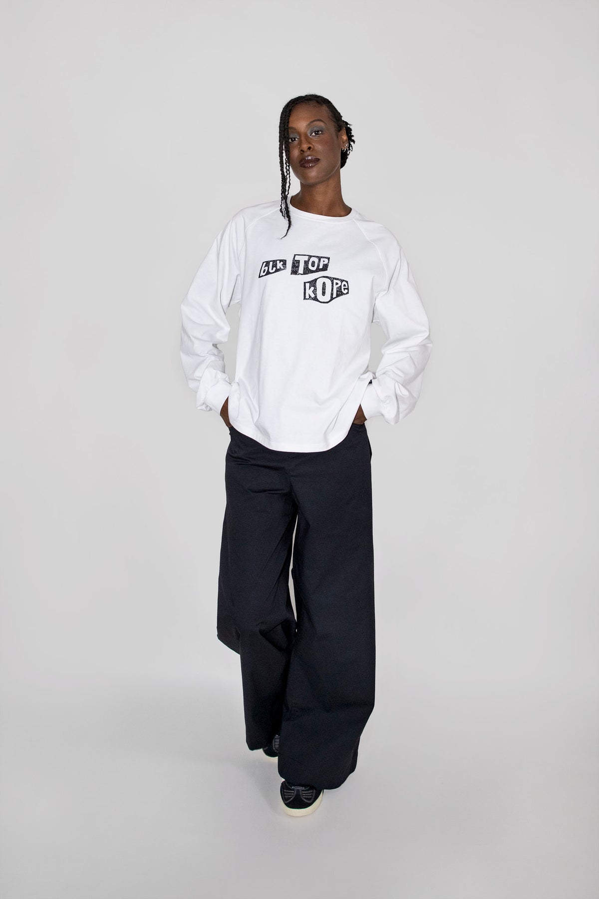 A model wearing a white long-sleeve sweatshirt with a black graphic print on the chest, paired with black wide-leg trousers, standing against a light gray studio background.